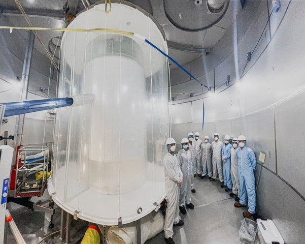 A group of people in white clean suits stand around a white cylindrical apparatus.