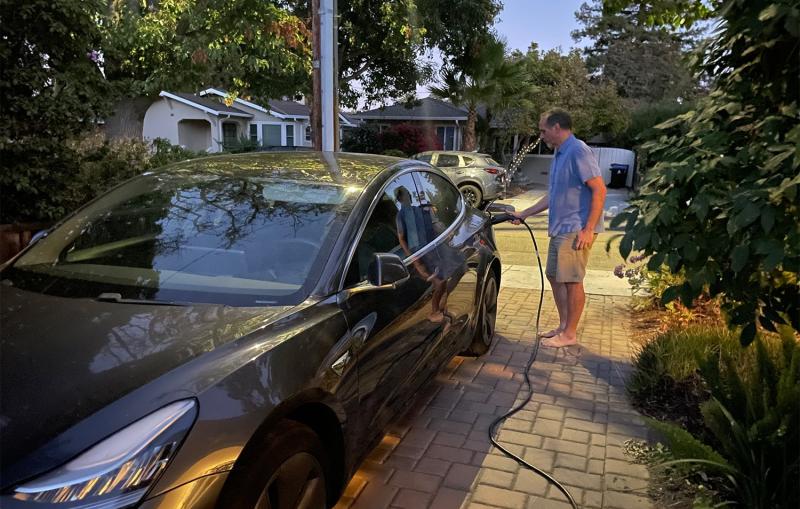 Photograph of a man plugging an electric cord into a gray car on a driveway.