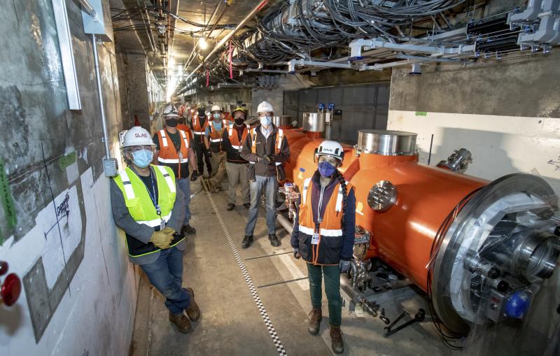 About a dozen people in safety vests, hard hats and masks pose in a concrete tunnel alongside a long red cylinder about 4-5 feet in diameter.