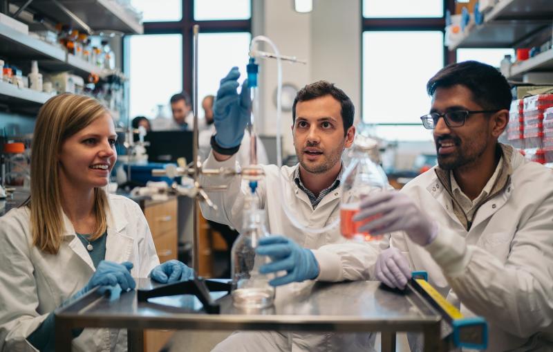 Three people in lab coats examine chemistry equipment.