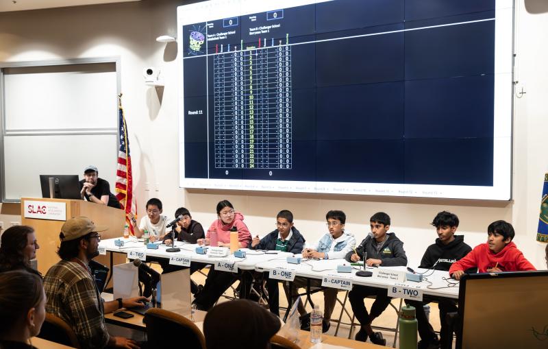 eight middle school students sit behind a long table at the front of an auditorium with buzzers in front of them as they compete in a science knowledge competition