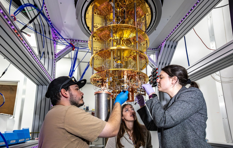 Researchers working with a dilution refrigerator, a piece of equipment that is made of gold-colored metal and resembles a chandelier.