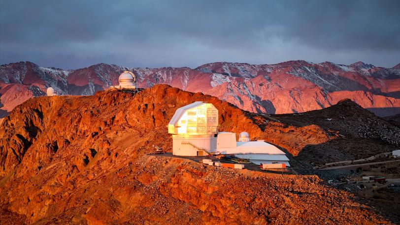 Aerial view of the Vera C. Rubin Observatory at sunrise, its dome glowing in warm light, surrounded by mountains.