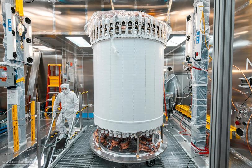 Large white cylindrical object suspended in clean room, person in white protective suit standing nearby, metallic walls surrounding.