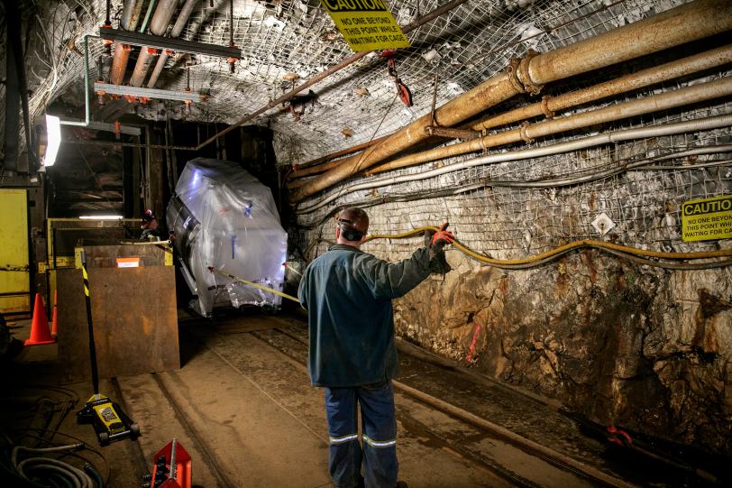 Underground tunnel with rocky walls, mesh ceiling with pipes, worker in safety gear directing movement of wrapped equipment moving through the tunnel.