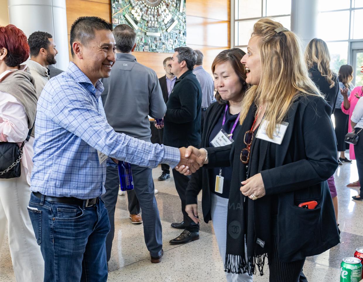 Two people greet each other with a handshake, smiling, surrounded by others.