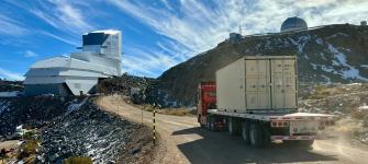 A truck carrying a shipping container down a gravel road nears a large telescope facility.