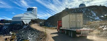 A truck carrying a shipping container down a gravel road nears a large telescope facility.