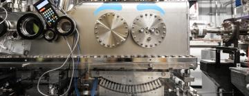 Large metallic machine in a lab, featuring valves, circular bolts, and digital displays with surrounding wires and tubing.