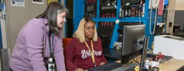 One woman standing, a second woman sitting down at a computer.