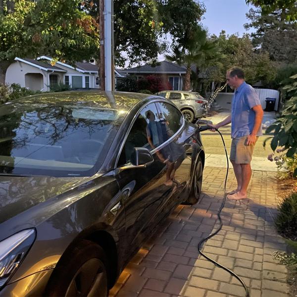 Photograph of a man plugging an electric cord into a gray car on a driveway.
