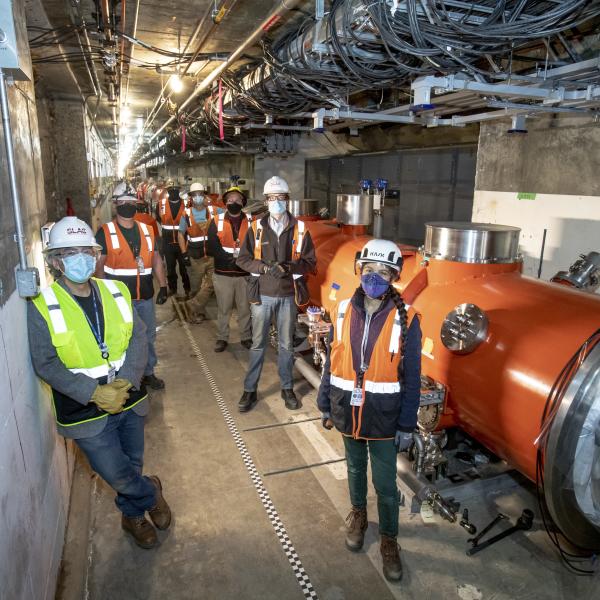 About a dozen people in safety vests, hard hats and masks pose in a concrete tunnel alongside a long red cylinder about 4-5 feet in diameter.
