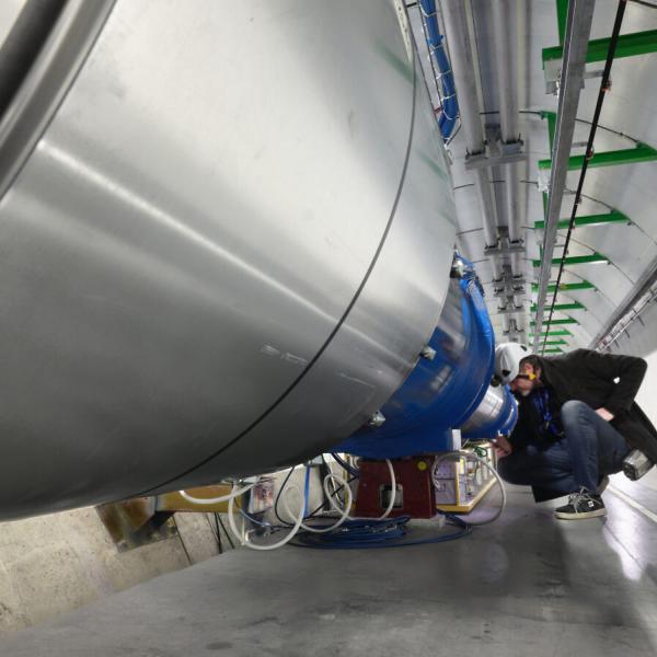 A person wearing a helmet kneels in front of a long metal particle accelerator tube.