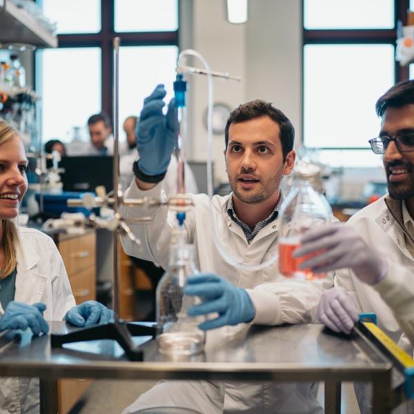 Three people in lab coats examine chemistry equipment.