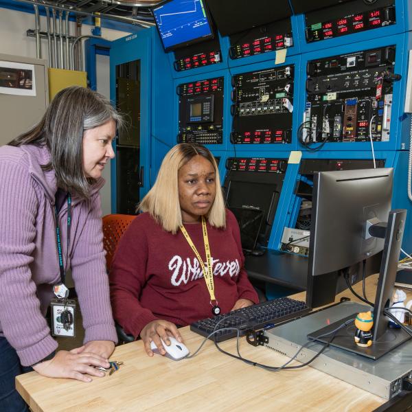 One woman standing, a second woman sitting down at a computer.