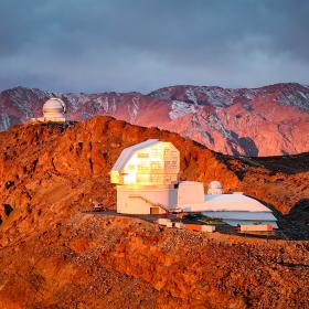 Aerial view of the Vera C. Rubin Observatory at sunrise, its dome glowing in warm light, surrounded by mountains.